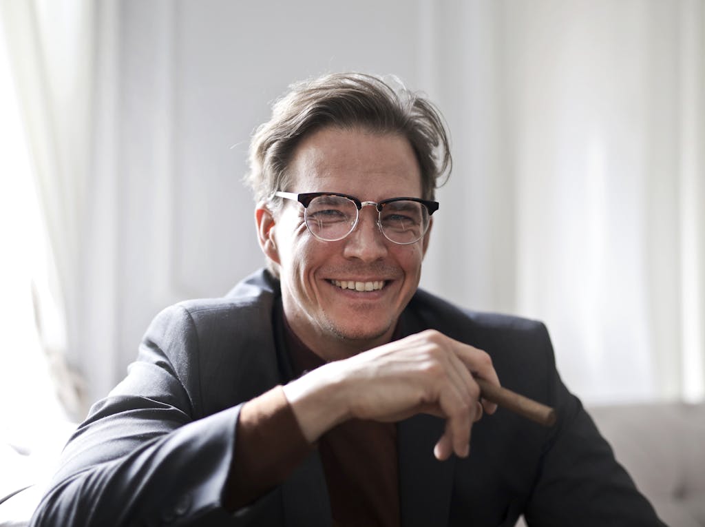 Cheerful businessman in glasses and suit holding a cigar, indoors.