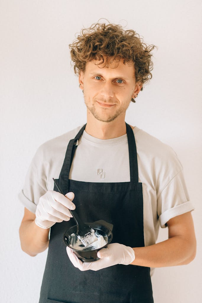 Smiling hairdresser holding dye brush and bowl, ready for a styling session in a salon.