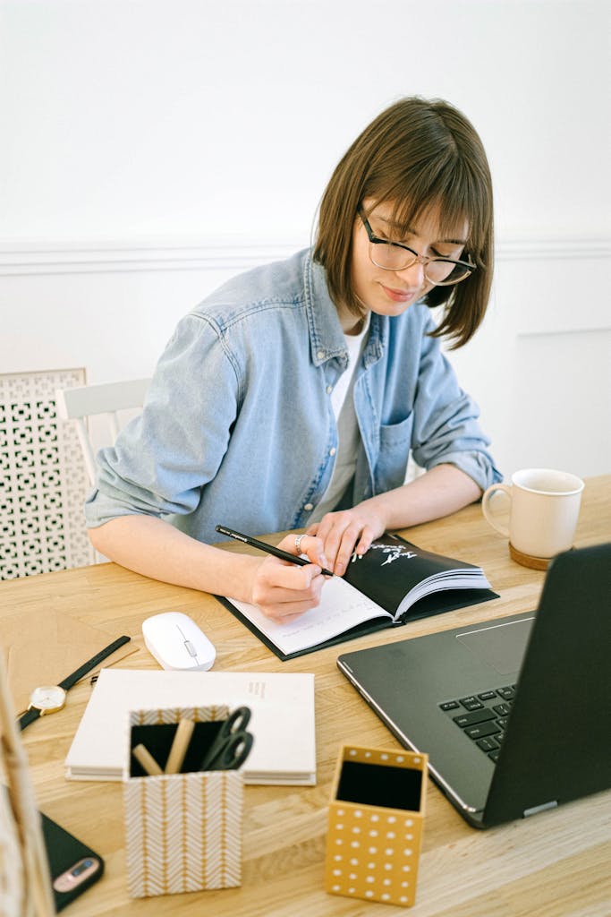Woman in glasses writing in notebook at home office desk with laptop and coffee.